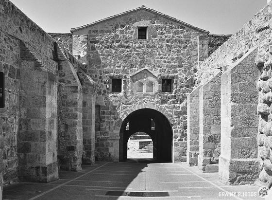 In a black and white image of the ancient Ciudad Rodrigo⁩ city gate with its arched entrance and rectangular windows. The pathway through the arch casts shadows, emphasizing the structure's rustic, historical appearance.