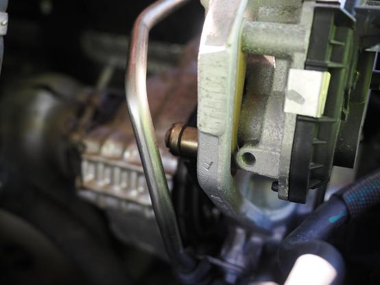 Detailaufnahme eines Motors. Im Vordergrund ist ein Metallteil mit einem daran geklippten Kunststoffteil. Links davon geht ein Bügel oder dünnes Rohr aus Metall vorbei. Rechts unten verschwindet ein Schlauch aus dem Bild, links ist im Hintergrund unscharf ein gewelltes Blech zu sehen.
