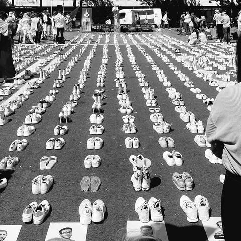 Rows of shoes are neatly arranged on the ground, each pair accompanied by a photograph placed in front of it. 

The scene are placed on Rotterdam to commemorate health care workers killed by Israel in Gaza.  

The image is in black and white, adding a somber tone to the scene.