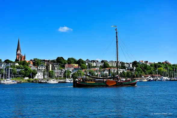 Auf der blauen Flensburger Förde gleitet ein Ewer in den Hafen. Die Segel sind nicht gesetzt, also fährt der alte Ewer unter Motor. Im Hintergrund sind die Häuser an Flensburgs Ostufer zu sehen, gekrönt von der Kirche St. Jürgen. Eine einzelne, kleine weiße Wolke ziert den strahlend blauen Himmel.