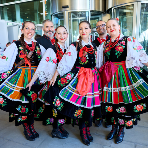 A group of men and women dressed in traditional Polish folk costumes pose cheerfully for a photo. The women wear colorful, embroidered dresses with striped skirts, floral patterns, white blouses with lace sleeves, and red bead necklaces. The men wear dark vests with red accents. They are standing in front of a modern glass building entrance.
