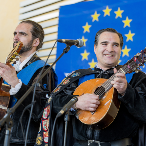 Two men in traditional Spanish student attire, known as 'tuna,' perform on stage with string instruments, one playing a guitar and the other a lute. They wear black capes adorned with colorful embroidered patches and blue sashes. A European Union flag with yellow stars on a blue background is visible in the background.
