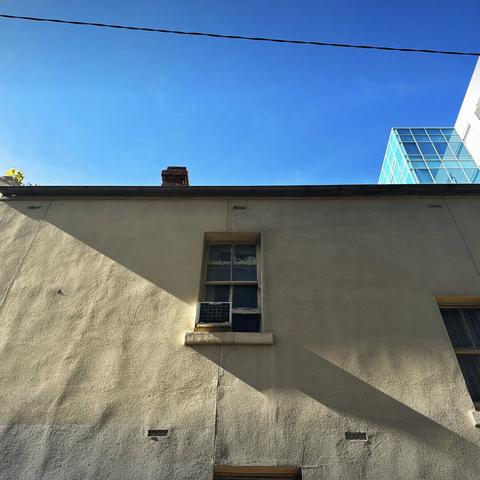 A diagonal shadow falls across an old building’s cream-coloured and uneven wall, across a single window frame containing 8 panes. A blue sky is above, as is a single wire.