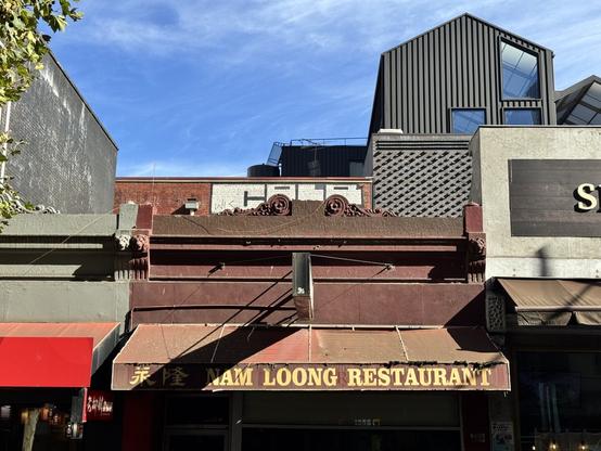 The shadow of a restaurant’s sign falls diagonally across its awning. The whole building is dark red, with the name “Nam Loong” in yellow. Beyond, a bluish sky and some newer buildings.