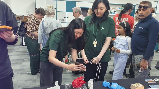 Two young volunteers in green t-shirts place a 3D printed heart onto Museum in a Box. One leans forward to photograph it with her Phone. In the back ground people are milling around and chatting.