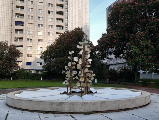 Im Vordergrund der Glasbrunnen, drei aufwärts strebende, an Blumen erinnernde Stelen mit Glasteilen. Im Hintergrund ein Hochhaus, Bäume rechts und links