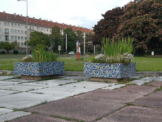 im Mittelpunkt des Bildes stehen zwei Pflanzkübel, die aus blau weißen Mosaiken gestaltet sind. Im Hintergrund ein langer Wohnblock, rechts im Hintergrund ein großer Baum