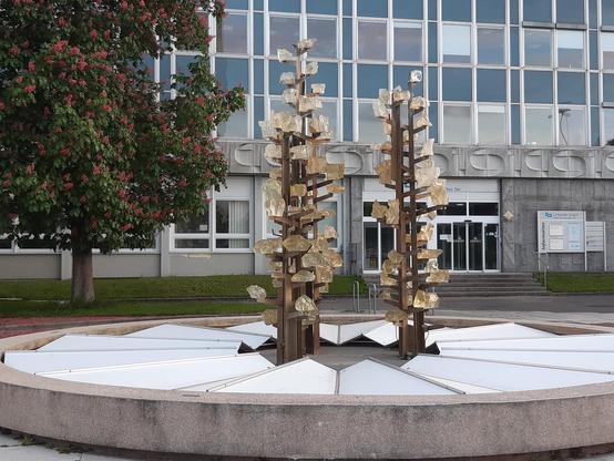 Im Vordergrund der Glasbrunnen, drei aufwärts strebende, an Blumen erinnernde Stelen mit Glasteilen. Im Hintergrund ein Haus mit blauer Fassade