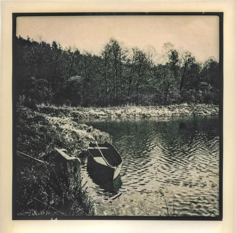 The image was printed using the lith print technique,  a photographic printing technique known for its distinctive high-contrast and grainy aesthetic, often with warm or sepia tones and dramatic shadow detail.

The image depicts a quiet, serene lakeside scene. In the foreground, a small rowboat is moored at the edge of the water, slightly tilted and partially reflected in the rippling surface. The shoreline is grassy and uneven, leading into a wooded area with tall, leafless or sparsely leafed trees, suggesting early spring. The dense forest in the background creates a dark, textured contrast to the lighter foreground. The overall mood is calm, nostalgic, and somewhat melancholic, enhanced by the lith printing process which gives the photograph a vintage, artistic look with rich blacks, soft highlights, and a warm tonal range.