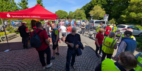 Auf einem Parkplatz ist ein roter Pavillion zum Sonnenschutz aufgebaut. Ein weißer Schriftzug besagt, dass er der Sparkasse Minden-Lübbecke gehört. Es stehen viele Menschen auf dem von Bäumen umrahmten Platz. Man sieht einige Fahrräder zwischen den Menschen.