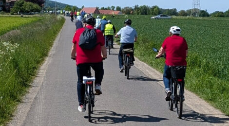 Man sieht viele Dutzend Menschen auf Fahrrädern auf einer schmalen asphaltierten Straße durch grüne Felder. Im Hintergrund ist ein bewaldeter Höhenzug zu sehen.