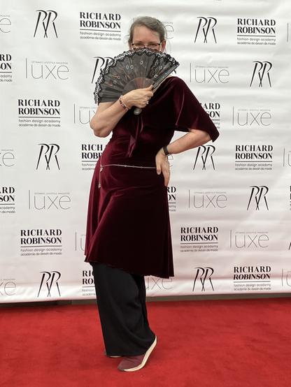 A woman wearing glasses and a burgundy velvet dress and black pants holds a black floral fan in front of her face , looking over it towards the camera. She stands on a red carpet in front of a banner that reads Richard Robinson Fashion Academy