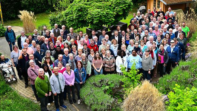 Das Bild zeigt die Konferenzmitglieder, die für ein Gruppenfoto posieren. Sie stehen im Freien, vor einem Garten mit Büschen und Bäumen. Im Hintergrund ist ein kleines Gartenhaus zu sehen. Die Menschen sind unterschiedlich alt und tragen verschiedene Kleidung.