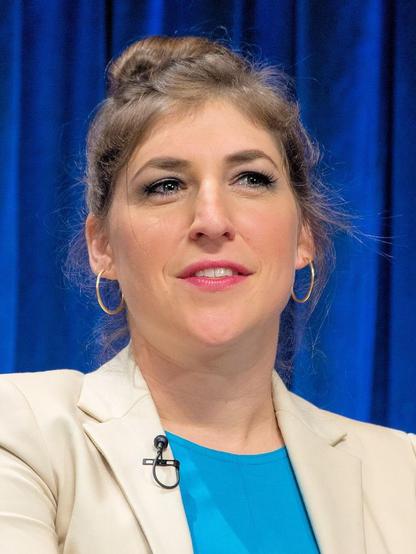 Ein close-up Porträt von Mayim Bialik. Sie trägt ein weißes Blazerjackett über einem blauen Oberteil. Die Schauspielerin trägt goldene Ohrringe. Der Hintergrund zeigt einen unscharfen blauen Vorhang, der eine Veranstaltungsumgebung andeutet.