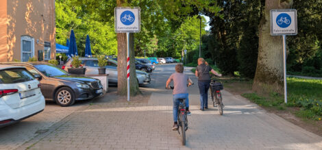 Eine Straße ist als reine Fahrradstraße mit Zeichen 244.1 gekennzeichnet. Eine Fraue schiebt ihr Rad zwischen den beiden Schildern hindurch, ein Junge fährt auf seinem Rad. Entlang des rechten Straßenrandes sind bis zum sichtbaren Ende der Straße KFZ quer zur Fahrtrichtung geparkt. Weiter hinten sieht man dichten Baumbestand, im Vordergrund rechts ist ein Bauwerk zu sehen.