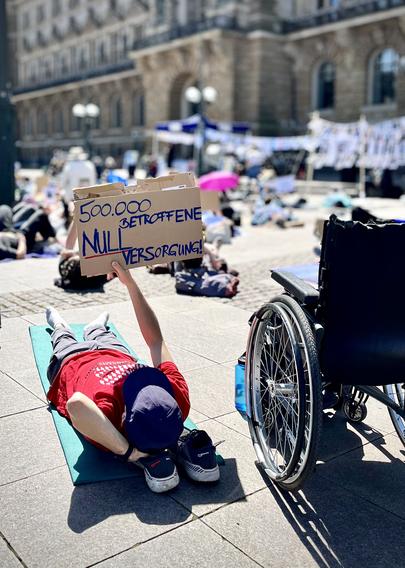 Rollstuhlfahrer liegt auf dem Boden, hält ein Schild hoch: „500.000 Betroffene, null Versorgung“