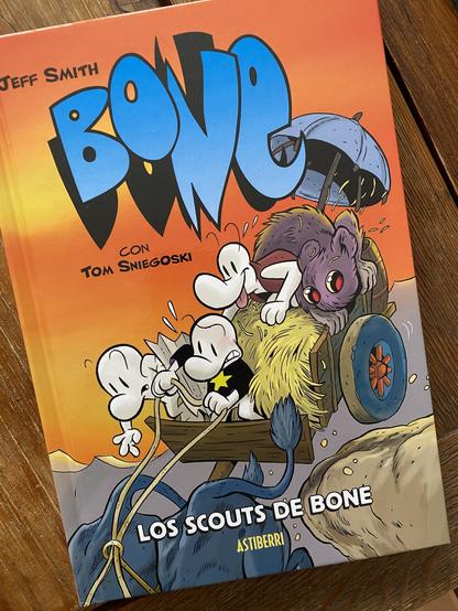 Portada de Los scouts de Bone. En ella se ve una ilustración en estilo cartoon de tres bones (unas criaturas blancas y narigudas) montados en un barro de bueyes cargado con paja. Al fondo se ve la monstrorrata que les acompaña (como su nombre da a sugerir, se trata de una rata monstruosamente grande. El carro está cayéndose por un acantilado y todos los personajes muestran cara de preocupación o miedo excepto el bone más alto que parece disfrutar de la aventura.