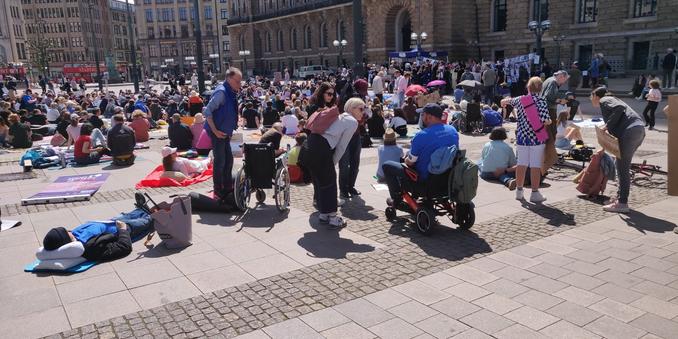 Foto von der Demo. Menschen liegen auf dem Hamburger Rathausmarkt und halten Schilder mit Fotos ihrer kranken Angehörigen hoch. Einige sind selbst betroffen und mit Rollstuhl gekommen.
