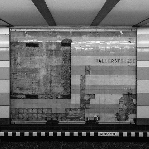black and white photograph taken in the hamburg u-bahn station hallerstrasse. the cladding has been partially removed, revealing the old tiles. sections of the tiles are missing, revealing the concrete. the station name is visibly faded on the old tiles.