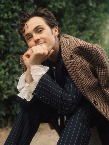 Misia Butler, actor blanco, moreno de pelo, lleva una chaqueta de tweed en los hombros, sentado posa sonriendo y apoyándose en sus manos.