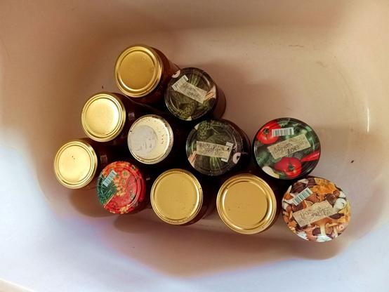 A plastic tub with large glass jars that you mostly see the metal lids of. The lids are shiny with some rust or patterns printed on them. A few have labels translating to "Plums, 2018".