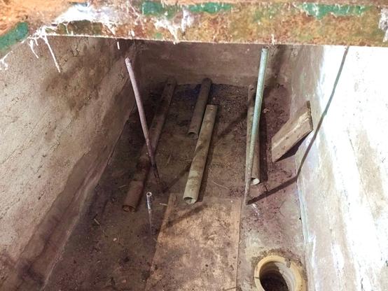 View down a trapdoor into a low retrofit basement with cement brick walls. There's a few small galvanized steel pipes cemented into the floor and a large concrete ring forming a sump. Four large drain pipes lay on the floor, next to them a wooden board. There's rust and spiderwebs on the frame of the trapdoor. It's somewhat dimly lit.