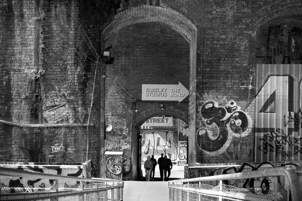 Looking through brick arches. A group of four people are walking through the arches towards the camera. Both the arches and the street visible through them have street art / graffiti on them.