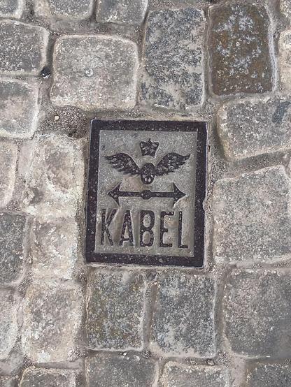 A slab between pavement tiles with iron engraving stating "cable" (in Polish). Above it, a bidirectional horizontal arrow, a symbol of spiked wheel with two wide wings emerging from it, and a crown above it.