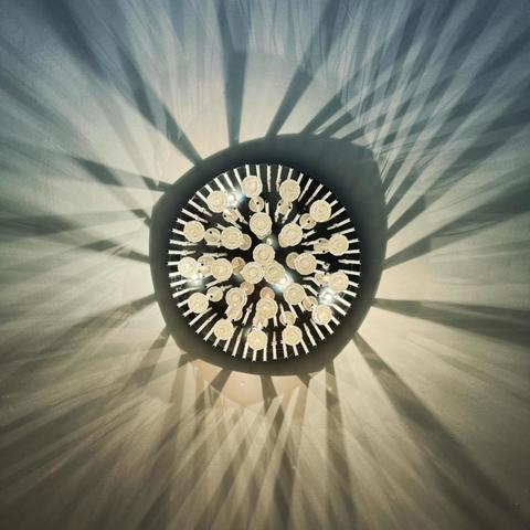 Das Bild zeigt einen Kronleuchter von unten, in dessen Mitte kreisförmig Lichtquellen angeordnet sind. Der Kronleuchter besteht aus mehreren Elementen aus Glas oder Kristall, die symmetrisch um einen Mittelpunkt angeordnet sind. Das Licht des Kronleuchters wirft komplexe Schatten an die Decke und bildet ein Muster aus scharfen, kantigen Linien, die vom Mittelpunkt ausgehen. Der Gesamteffekt ist ein dynamisches Wechselspiel aus Licht und Schatten.