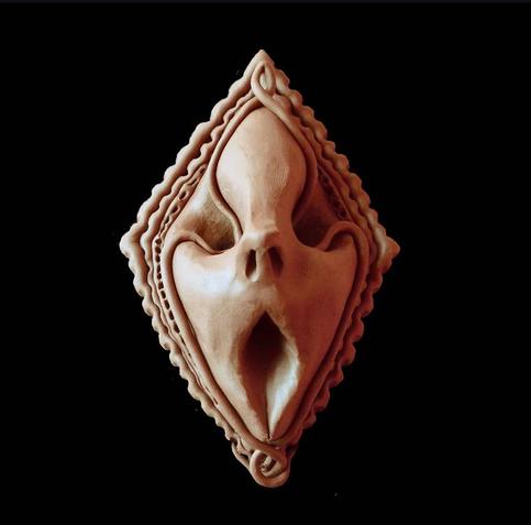 The image shows a diamond-shaped clay or ceramic sculpture with facial features resembling a mask. The sculpture has a flesh-toned, light beige color and features abstract, stylized facial elements including what appears to be an elongated mouth opening in the center, nostril-like indentations above, and subtle eye-like forms. The piece has decorative scalloped or rippled edges around its diamond perimeter with small curled details at the bottom. The artwork is photographed against a solid black background, creating a strong contrast that highlights the textural details and organic contours of the sculptural form.