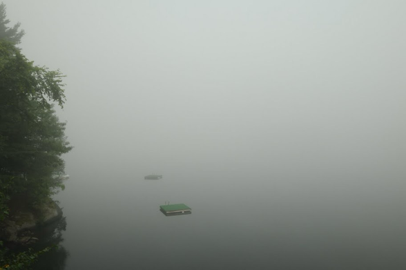 A foggy scene of a lake with some evergreen trees at right and a couple of square, floating dive platforms on the water. The lake itself is barely perceptible due to the fog.