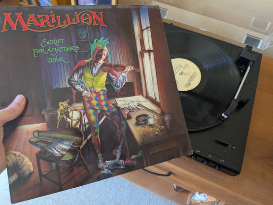 A photograph of a modern vinyl record player and me holding "Script for a Jesters Tear" by Marillion in the foreground.