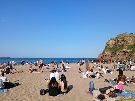 A very sunny King Edward's Bay at Tynemouth. The beach is very busy, and the sky and sea are blue. To the right you can see the cliffs