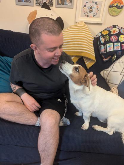 A white transmasc sat on a blue sofa with a white and brown jack russel. The human is wearing a grey t shirt, blue shorts, and sports socks. The human and dog are looking at each other