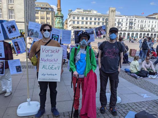Drei Personen tragen Atemschutzmasken und stehen auf einem belebten Platz. Eine davon hält ein  Schild in der Hand, auf welchem „Vorsicht statt Nachsicht - ALGE e.V.“ und „www.alge-hamburg.de“ steht.