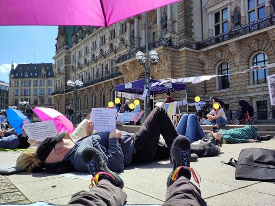 Liegenddemo für Betroffene von ME/CFS auf dem Hamburger Rathausmarkt. Mehrere Menschen liegen auf dem Rücken und halten Schilder mit Forderungen and Politik und Medizin. Im Hintergrund sieht man einen Infostand. Es ist sehr sonnig.
