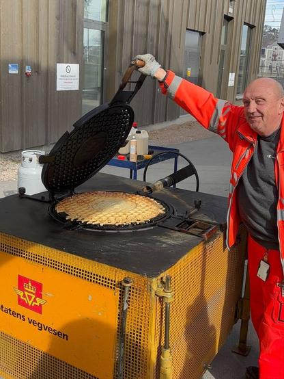 A man wearing red high-vis road worker jacket and pants is holding the mentioned waffle iron open. He's holding a handle that's been welded to a manhole cover, which is hinged into a base on the other side. The base is a big yellow box with a logo and a blackish top. On the top of the base is another manhole cover -- the bottom plate of the iron.

A finished waffle taking up almost the entire area of the manhole cover is presented on top of the bottom plate.