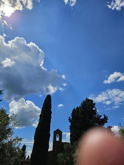 Il campanile di un oratorio di campagna è in mezzo a due cipressi. Il cielo è azzurro con nuvole bianche. Nell'angolo in basso a destra c'è un pezzo del mio indice fuori fuoco. Foto controluce.