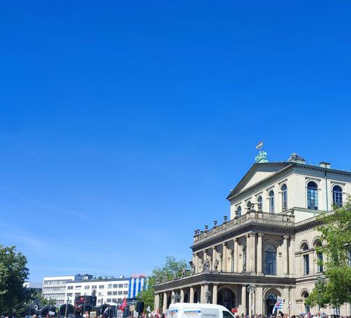 Demonstranten für AFD-Verbot vor dem Opernhaus Hannover, auf dem eine Regenbogenflagge unter strahlend blauem Hinmel weht.