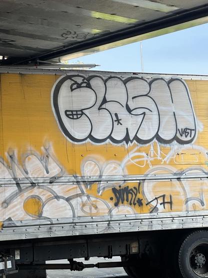 FLASH in white bubble letters with a black outline on a yellow truck, surrounded by additional miscellaneous and faded black and white graffiti. the F is a face, there's an asterisk in the lowercase A, and the letters VST are in the bottom of the H.