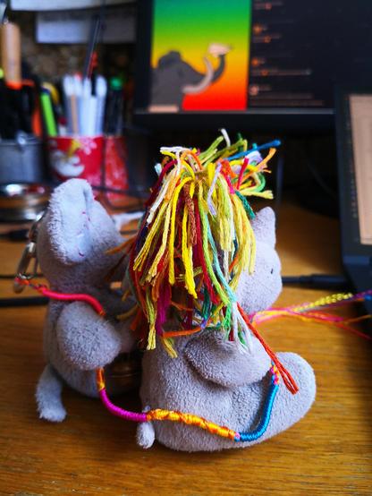 Die beiden kleinen Plüschies Elefanty Elfi und Einhorn Carola sitzen auf dem Schreibtisch neben dem Computer.
Das ganze Foto ist sehr bunt und farbintensiv:
Carolas Regenbogenhaare leuchten, Elfi hat ihr Ersatzband um Carola geschwungen, im Hintergrund ist auf einem zweiten, sehr großen Bildschirm die erweiterte Weboberfläche von Mastodon zu erkennen, deren linke Spalte mittels CSS so modifiziert wurde, dass hinter der Mastodon-Zeichnung (Kopf im Profil mit Papierflieger im Rüssel) einen Farbverlauf in Regenbogenfarben zu erkennen ist.