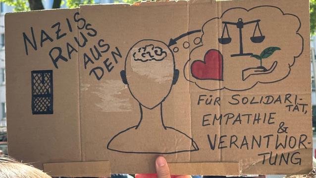 Schrift auf dem Plakat: Nazis raus aus den …
es folgt ein gemalter Kopf mit stilisiertem Hirn. Rechts davon geht es weiter: Für Solidarität & Vielfalt & Verantwortung. 
In einer Gedankenblase sind ein Herz, eine Waage und eine Hand auf der eine Pflanze wächst. Ein Pfeil zeigt von da zum Gehirn.