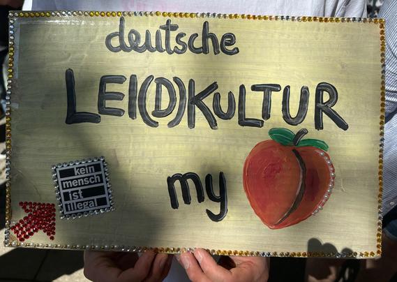 Ein weißes Plakat umrahmt mit orangen und weißen Glitzersteinchen: Deutsche Leidkultur my ass. Das D in Leidkultur ist eingeklammert. Statt dem Wort ass ist ein Pfirsich aufgemalt. Unten links ein Sticker: Kein Mensch ist illegal. Ein Pfeil aus roten Glitzersteinchen zeigt darauf.