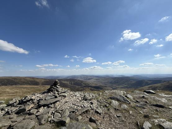 A rocky mountain summit features a stacked stone cairn in the foreground, with a panoramic view of rolling hills and valleys under a blue sky adorned with fluffy clouds.