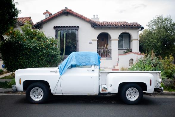 A color photograph of a white ‘70s Chevy side step truck with a blue tarp over the windshield, in front of a white Spanish architecture house