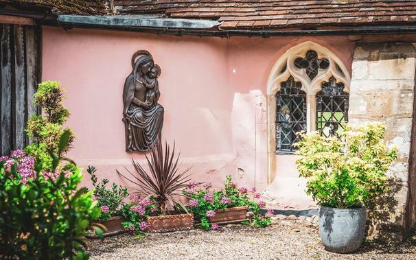 Stone relief of a seated Virgin Mary with the Christ-child on her knee, against pink-washed walls in a sunny medieval courtyard garden at Clare Priory in Suffolk.