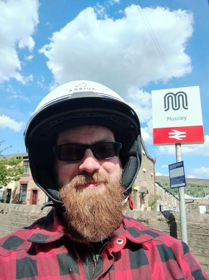 A selfie of a smiling bearded man in a red and back checked jacket and wearing an opennface motorbike helmet. He's in front of the Mossley railway station sign.
