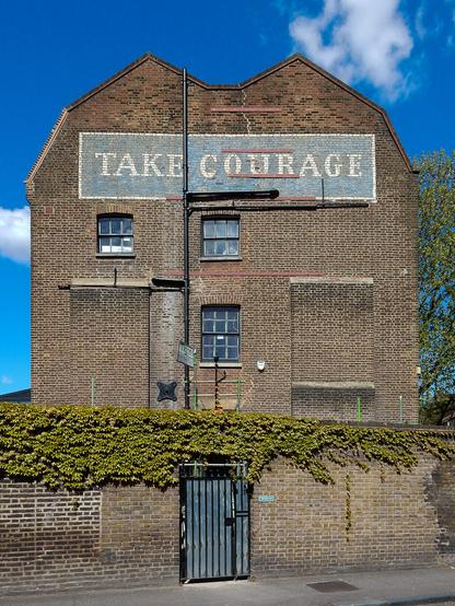 Photo of the side of a brick detached house which has a painted sign across the width of the upper portion of the wall. The sign says "Take Courage".