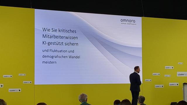Man sieht aus einem Publikum heraus auf die Präsentation eines Edu-IT-Dienstleisters in einem zitronengelben CI-Look. Ein männlicher Präsenter im Anzug steht auf der Bühne, der Screen hinter ihm sagt: "Wie Sie kritisches Mitarbeiterwissen KI-gestützt sichern und Fluktuation und demographischen wandel meistern"
