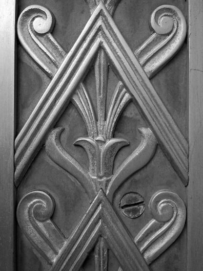 Black and white close-up of a decorative metal panel which features stylized plant forms overlaid with chevrons